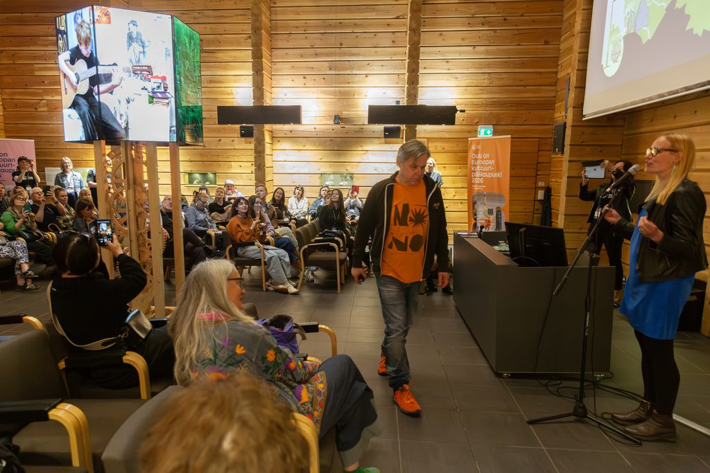 Joukko ihmisiä hirsirakenteisessa salissa, vasemmalla suuri kello, jonka päällä videonäyttöjä.