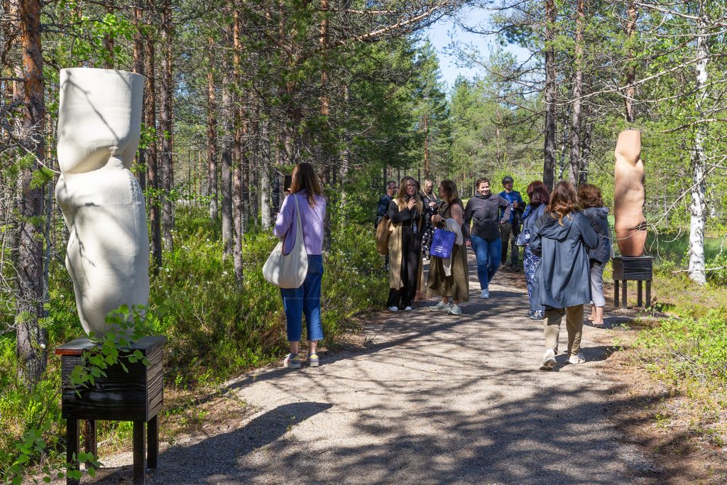 Joukko ihmisiä kulkee metsäpolulla, jonka laitamilla saviveistoksia.
