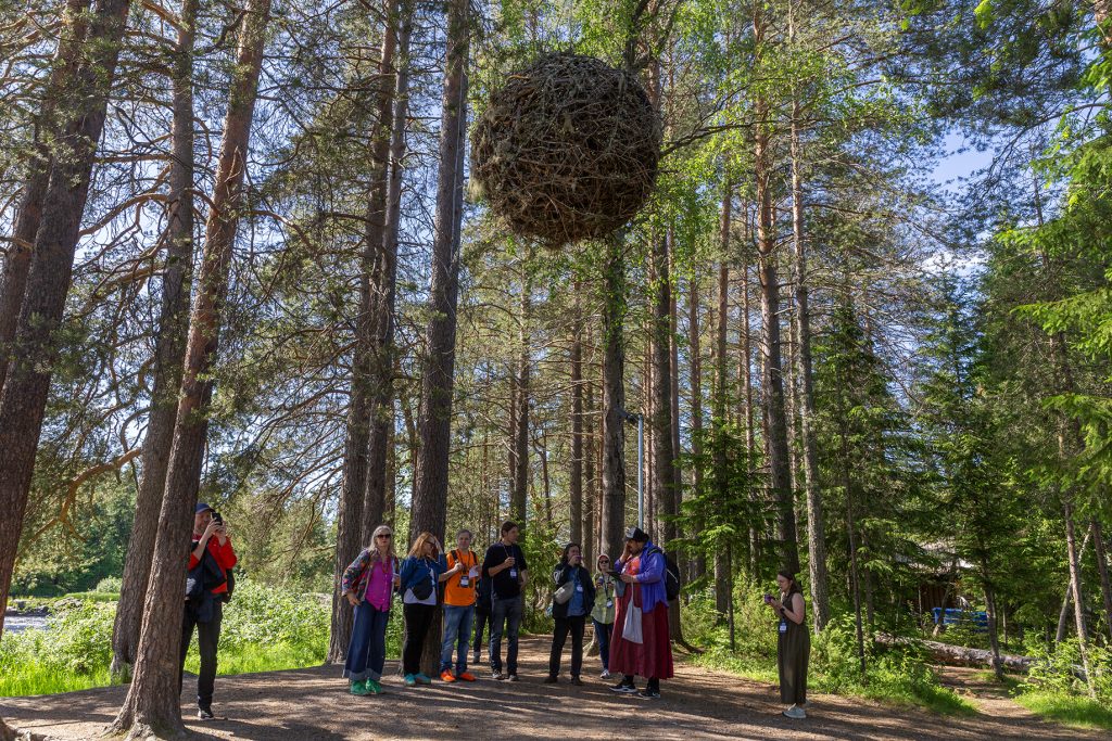 Joukko ihmisiä puiden keskellä, ylhäällä roikkuu katajista ja jäkälistä koottu suurehko pallo.