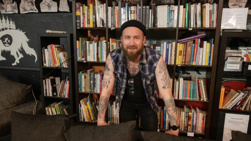 Parrakas, tatuoitu mies musta pipo päässään nojaa edessään olevaan sohvaan, taustalla kirjahylly täynnä kirjoja.