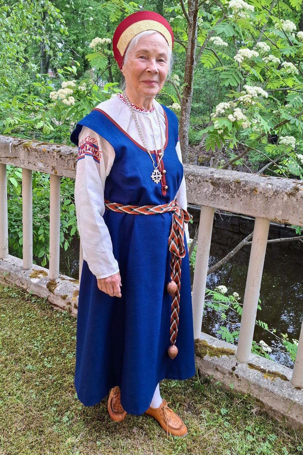 Kansallispukuinen nainen seisoo vanhan puuaidan vieressä, taustalla pensaita.