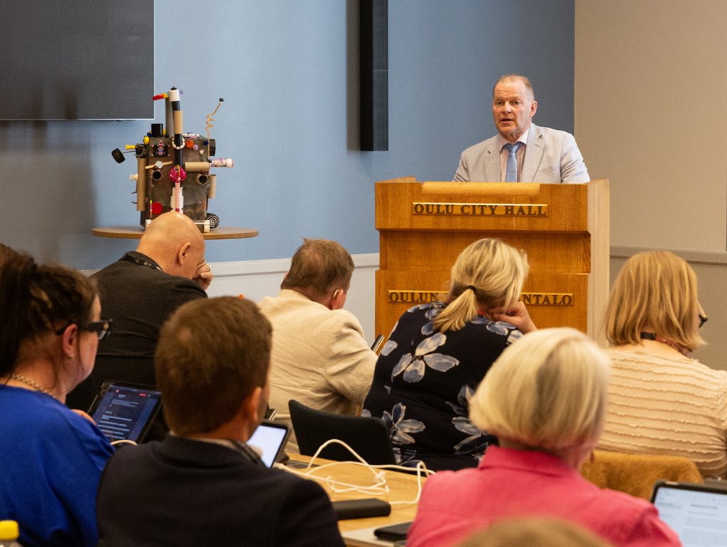 Mies puhuu valtuustosalissa, vasemmalla lasten tekemä robottimainen hahmo.