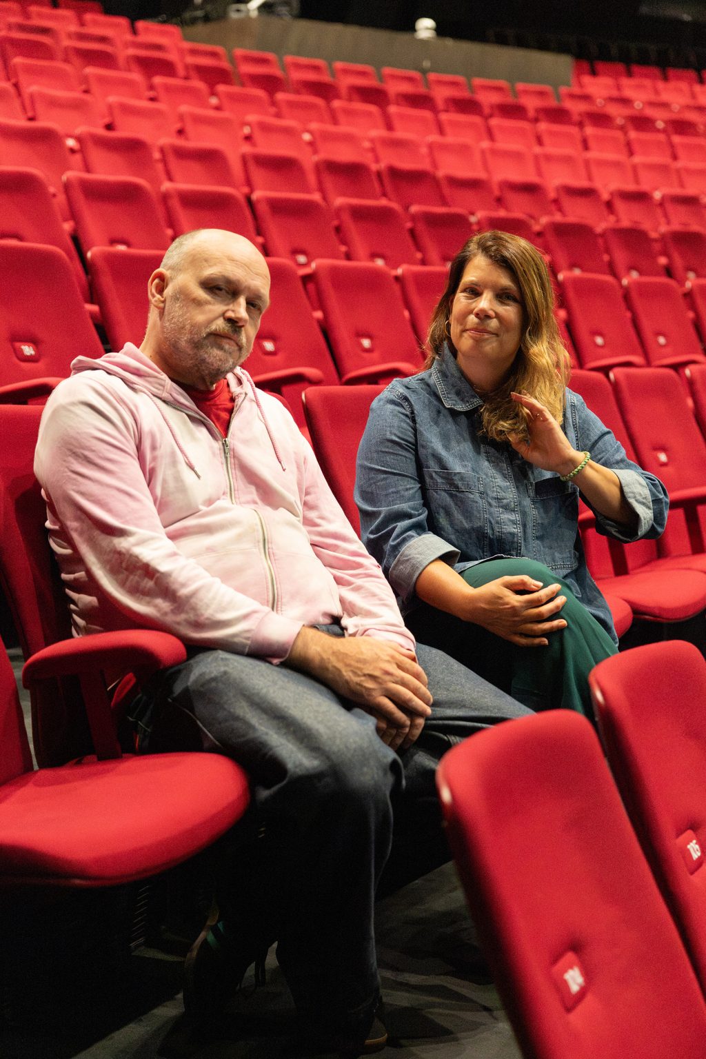 Kalju mies ja ruskeahiuksinen nainen, molemmat keski-ikäisiä, istuvat tyhjän teatterin katsomon punaisilla istuimilla.