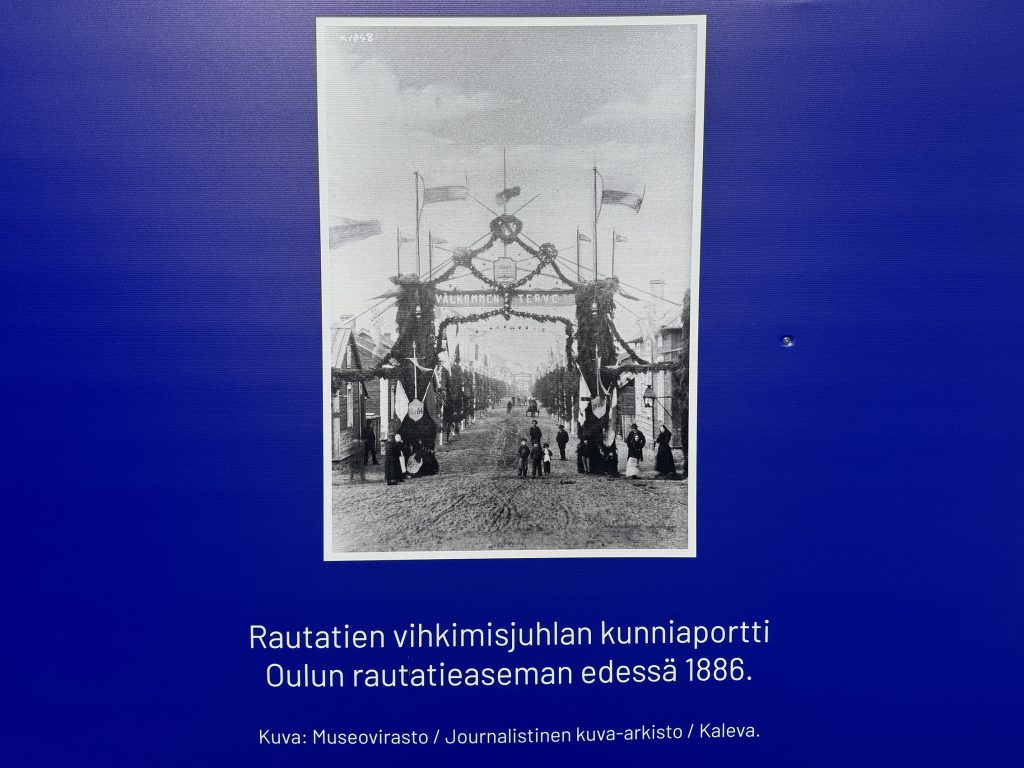 Siniselle taustalle painettu mustavalkokuva 1800-luvun asemanseudusta.