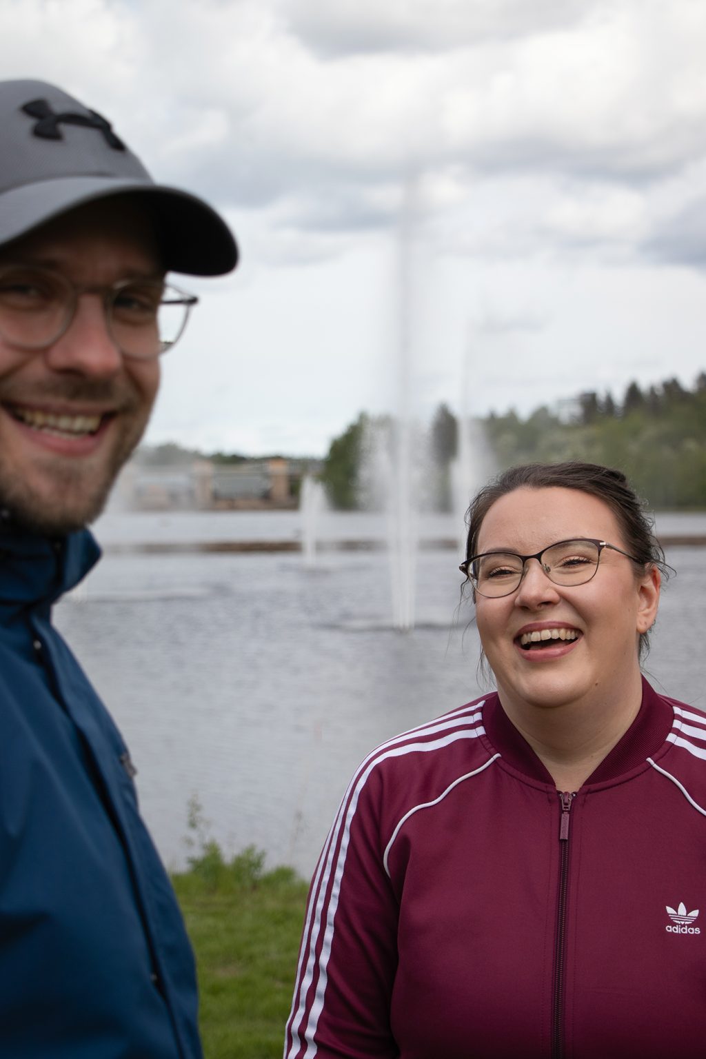 Verryttelyasuihin pukeutuneet mies ja nainen hymyilevät puistossa, taustalla suihkulähde vedessä.