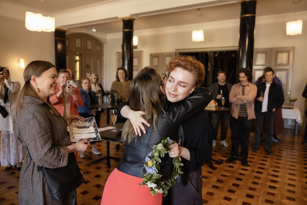 Nainen halaa juhlasalin aulassa nuorta miestä, jolla kukkaseppele kädessään, vasemmalla nainen kunniankirja kädessään, taustalla joukko muita ihmisiä.