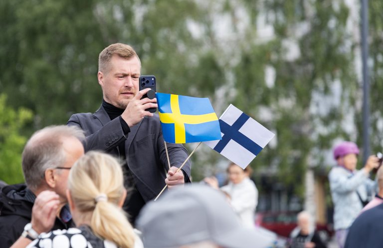 Vaalea mies kuvaa puhelimellaan väkijoukon keskellä, kädessään Ruotsin ja Suomen pienoisliput.