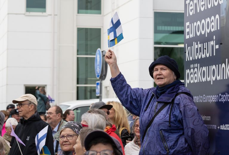 Iäkäs nainen heiluttaa Suomen ja Ruotsin pienoislippuja väkijoukon takana.