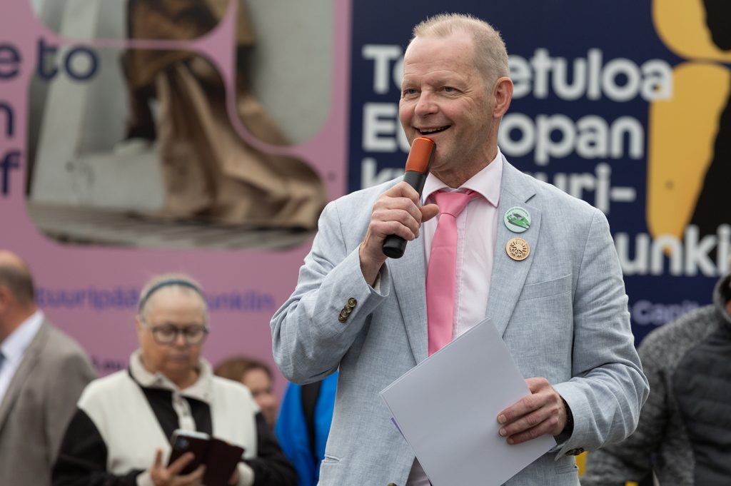 Mies vaaleassa pikkutakissa puhuu yleisön keskellä.