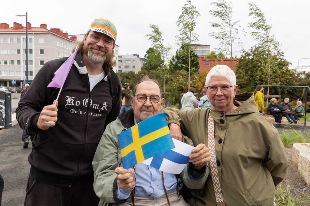 Kaksi miestä ja nainen poseeraavat ulkona, vanhempi miehistä istuu keskellä ruotsin ja suomen pienoisliput käsissään.