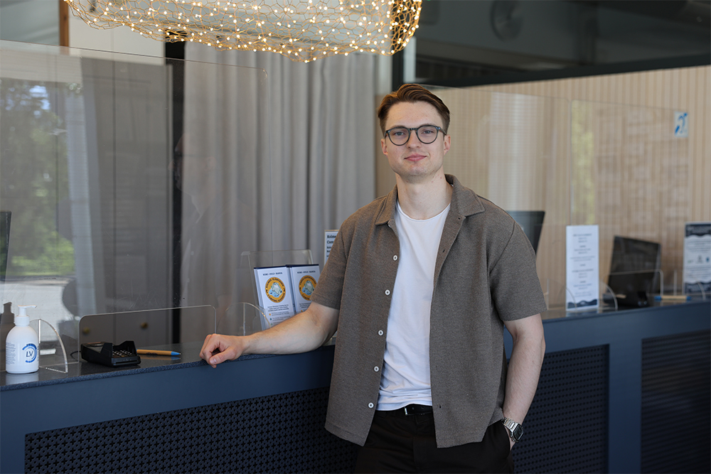 Nuori, silmälasipäinen mies nojaa majoitusliikkeen vastaanottotiskiin.