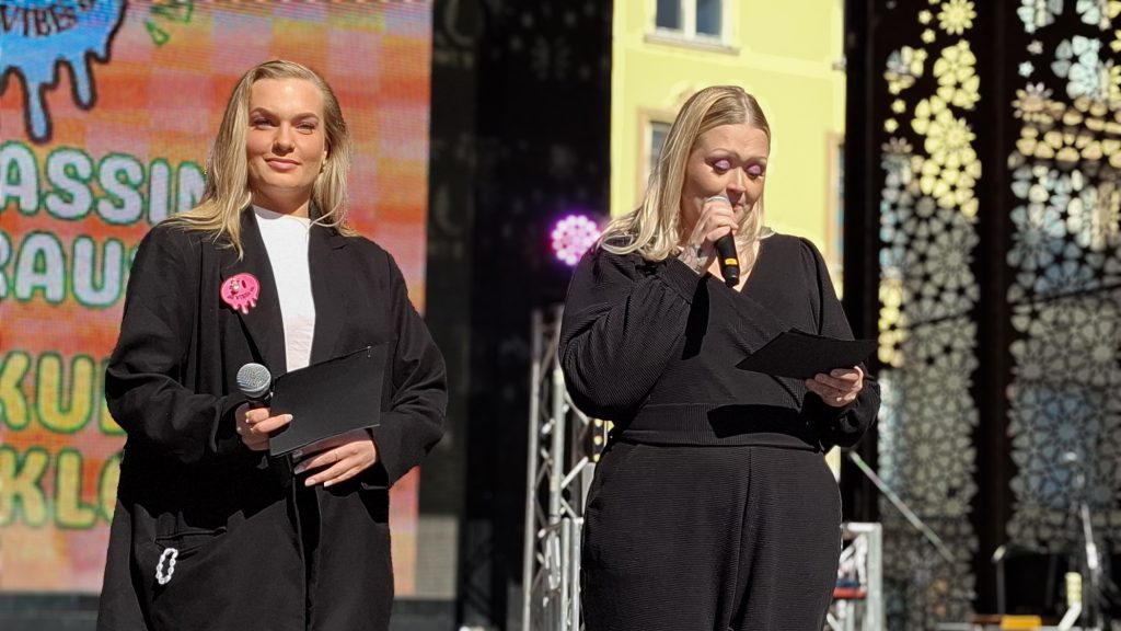 Kaksi nuorta, vaaleaa naista lavalla, toinen puhuu mikrofoniin.