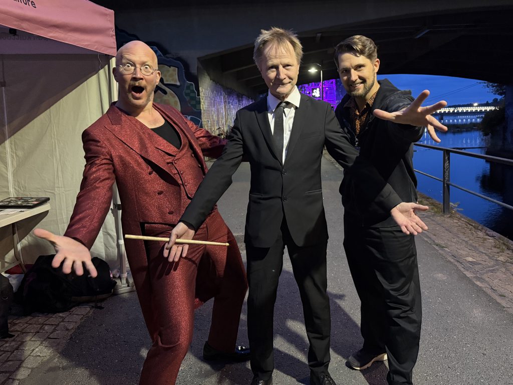 Kolme miestä poseeraa kädet levällään pimeässä illassa.