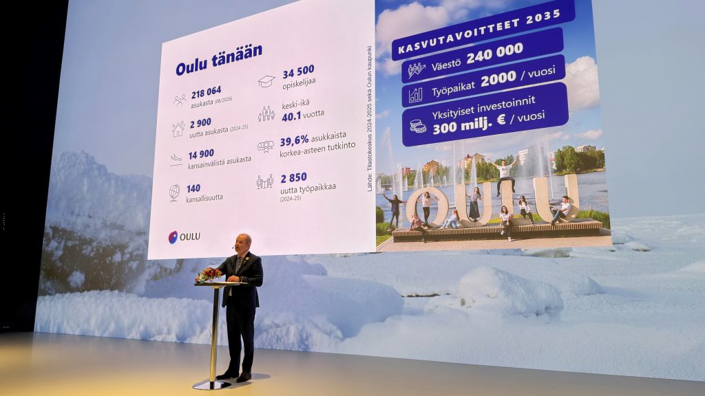 Kaupunginjohtaja puhuu, taustalla seinän kokoinen näyttö, jossa lukuja ja kuvia.