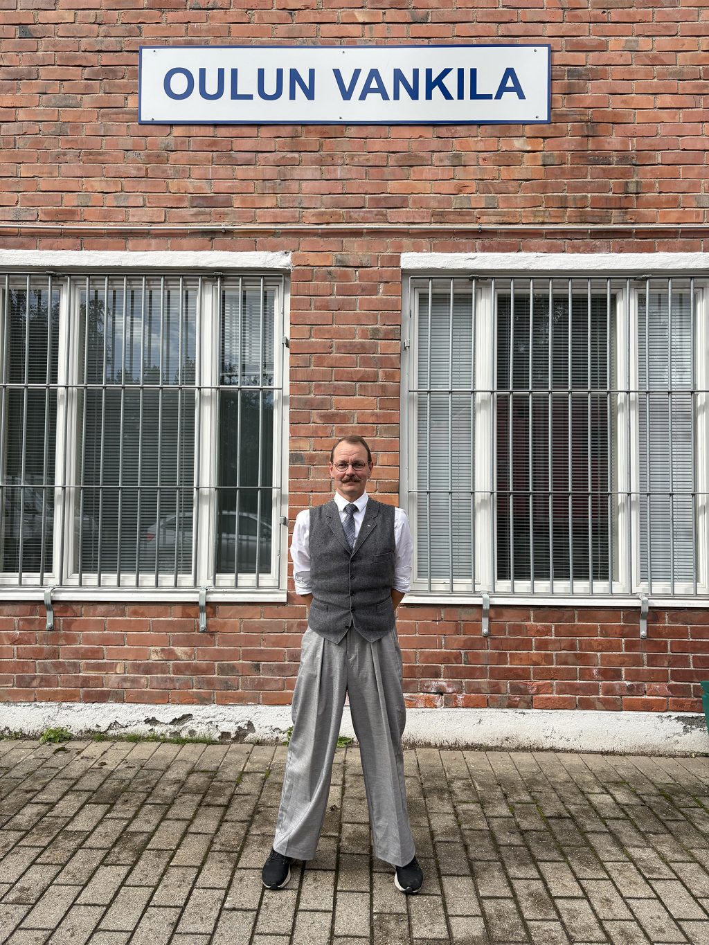 Liiviin ja solmioon pukeutunut mies poseeraa kädet selkänsä takana, taustalla tiilirakennuksen seinässä kyltti "Oulun vankila"