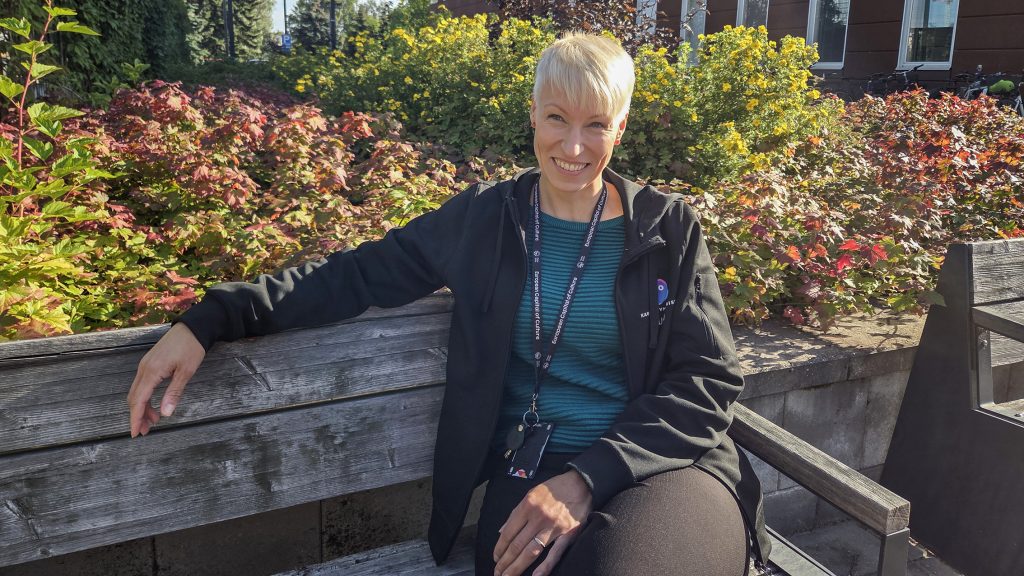 Vaalea, lyhyttukkainen nainen hymyilee penkillä istuen, taustalla pensaita.