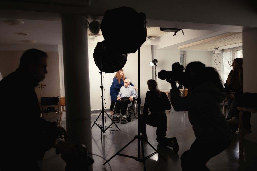 Joukko ihmisiä valokuvausstudiossa, kuvattavana istuu iäkäs mies.