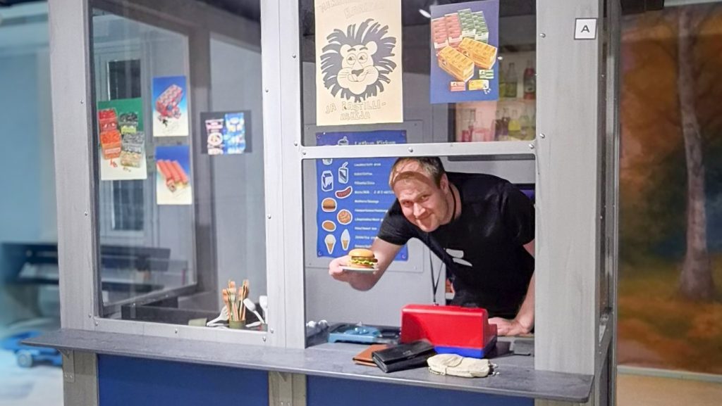 Mies kurkottaa lavastetun kioskin ikkunaluukusta hymyillen.